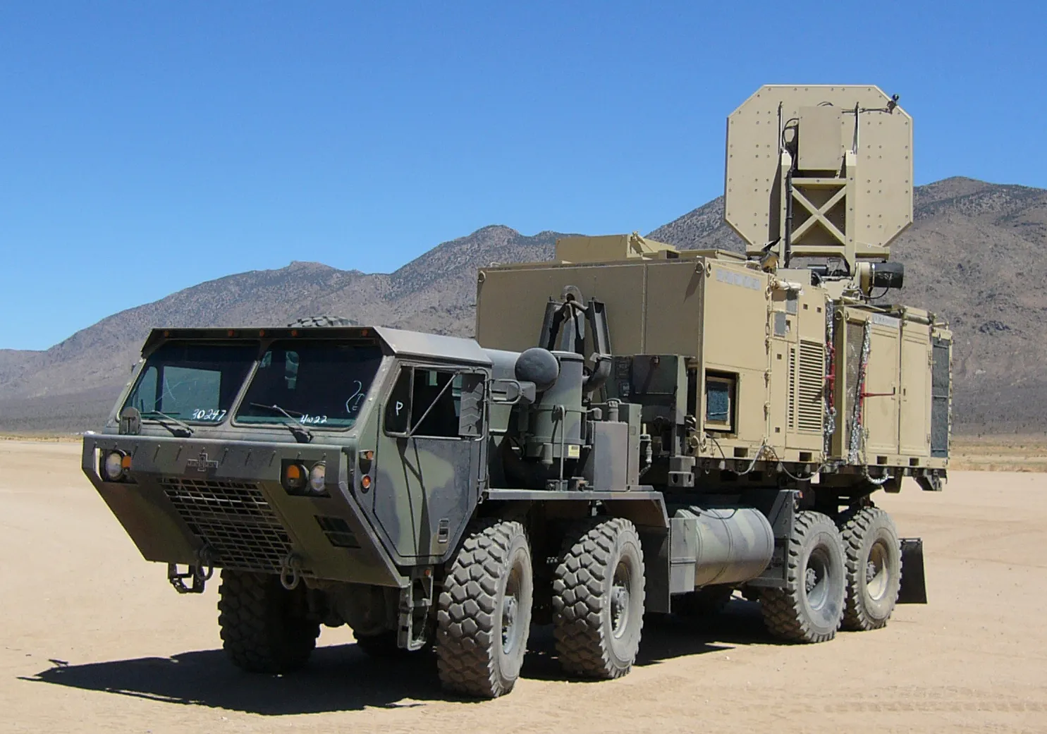 A large flat antenna dish mounted on the back of a tan military truck in the desert.