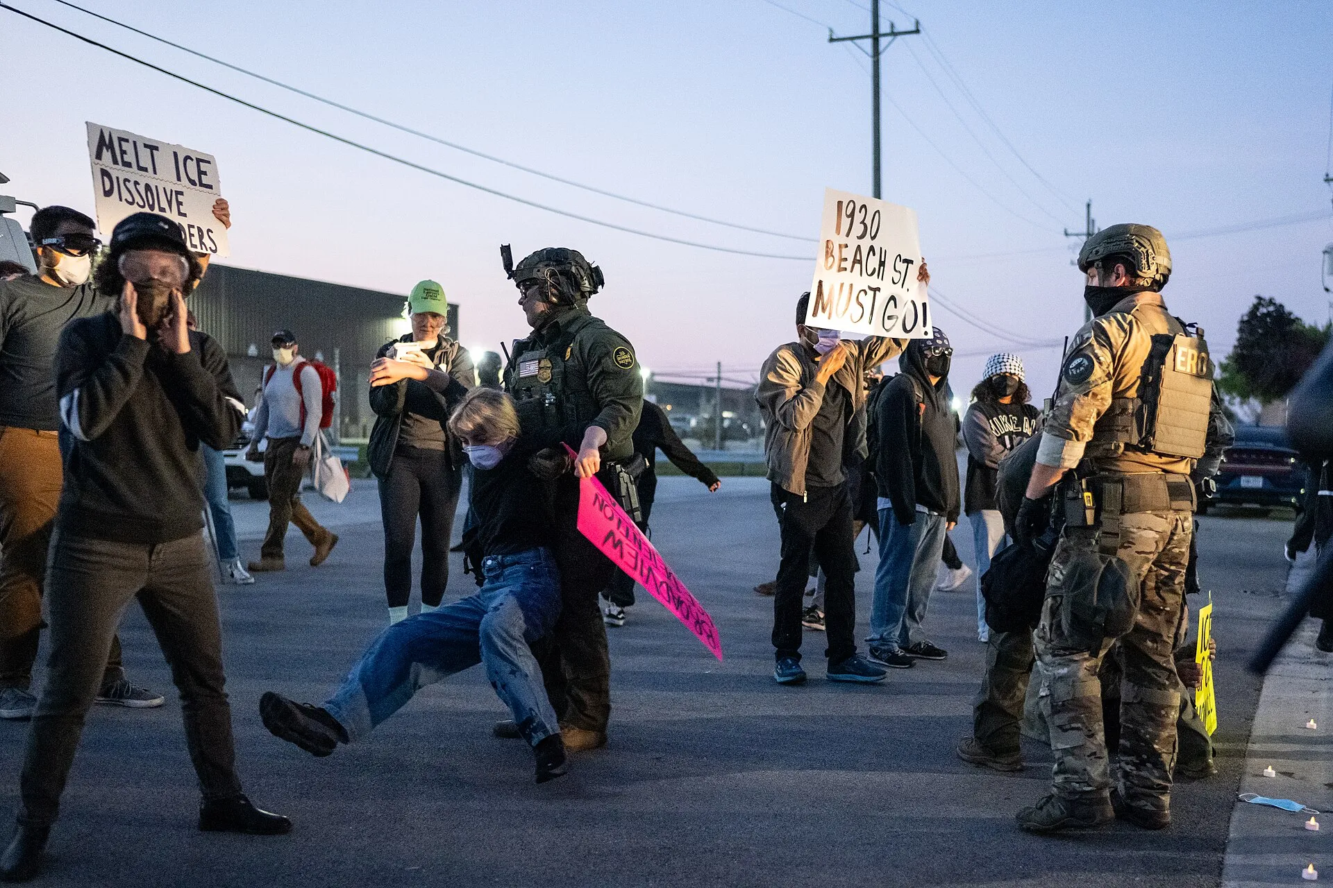 Federal agents in tactical gear outside the Broadview processing center; one pulls a protester to the ground while others hold signs.