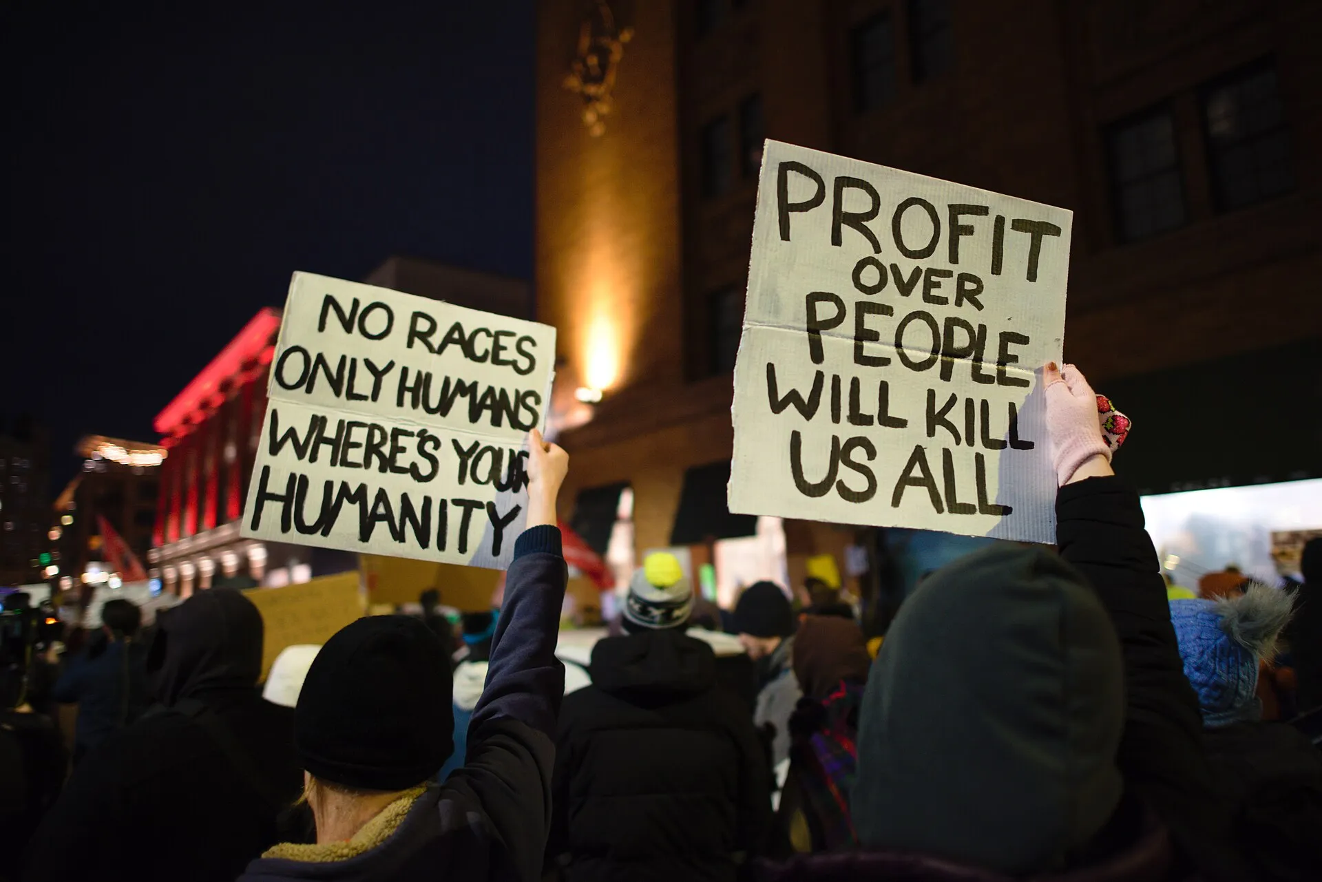 A crowd at night in downtown Minneapolis holding hand-lettered signs, one reading 'Profit over people will kill us all.'